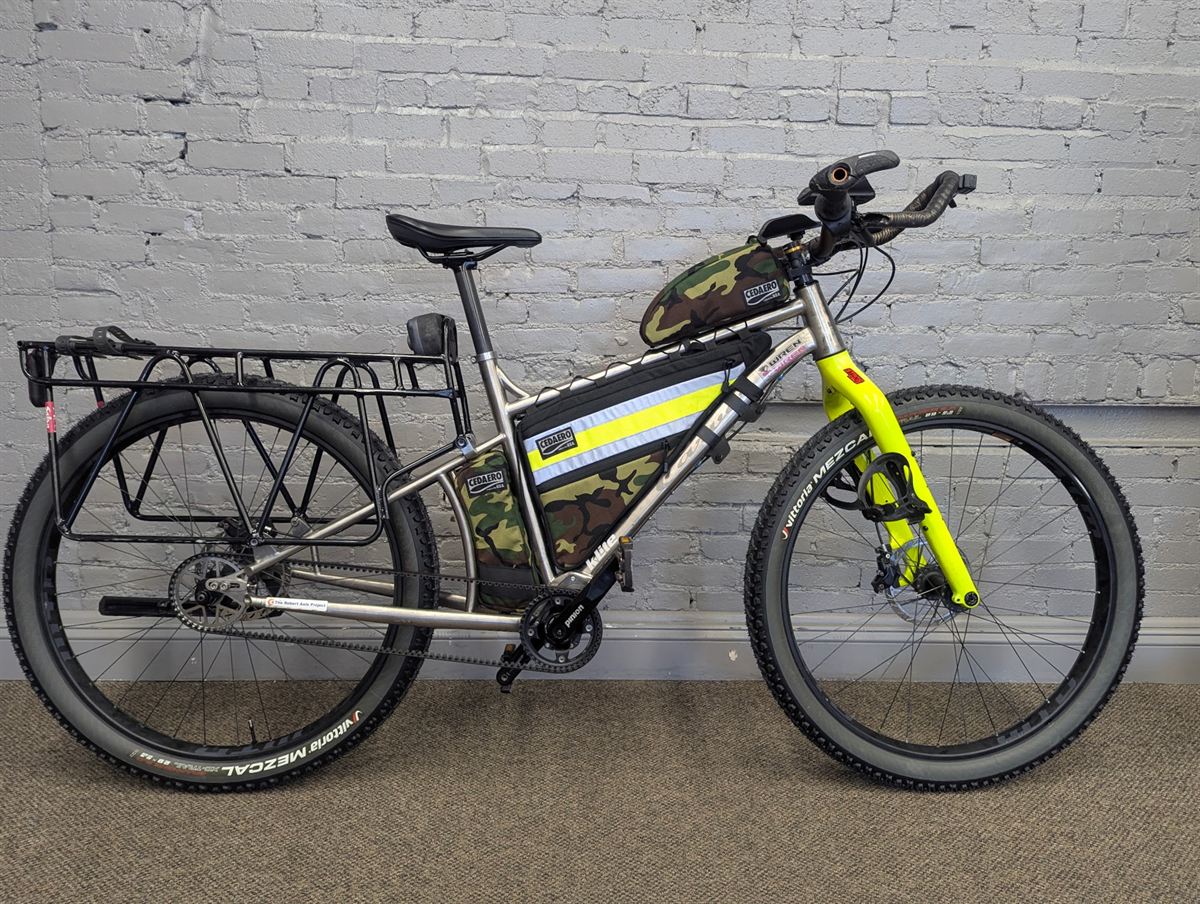 Titanium fat-bike frame set up for summer on 29x2.8 knobbly tyres, side on against a white-painted brick wall, showing the Pinion gearbox at the cranks and the belt drive running back to the hub.
