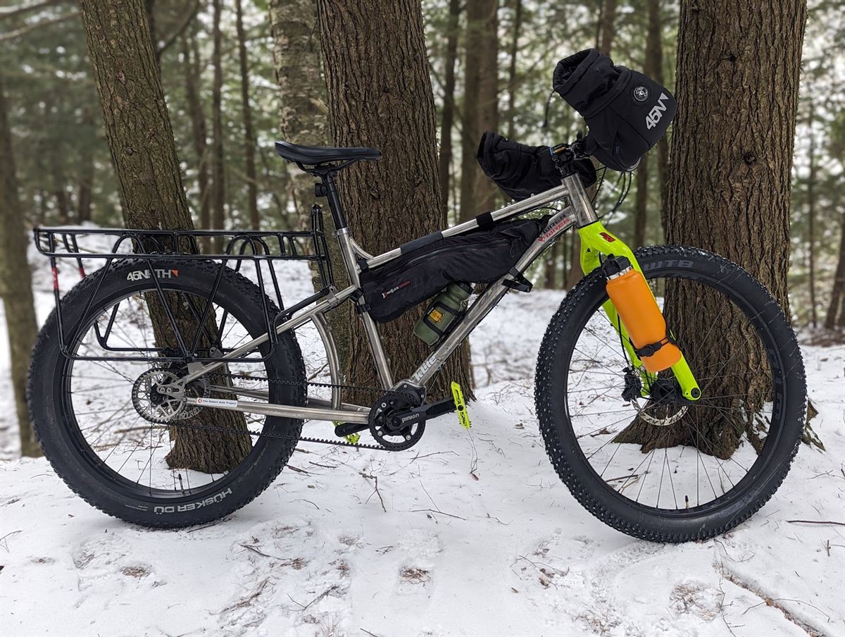 The same titanium bike in snow between pine trees, now on 27.5x4.5 studded winter tyres with a framebag, handlebar pogies and a rear rack.