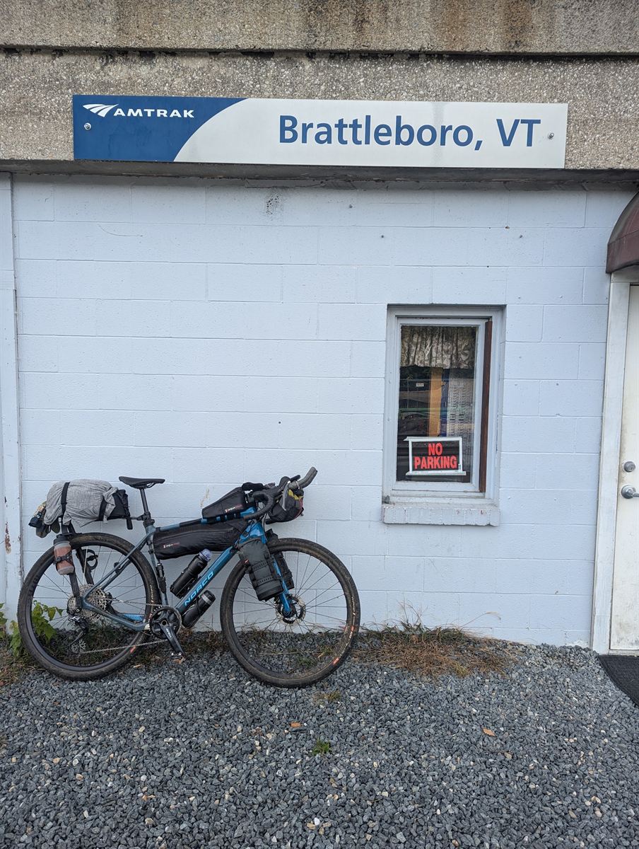 Blue Norco gravel bike loaded with bikepacking bags, leaning against the white cinderblock wall of the Amtrak station at Brattleboro, Vermont, under the station sign.
