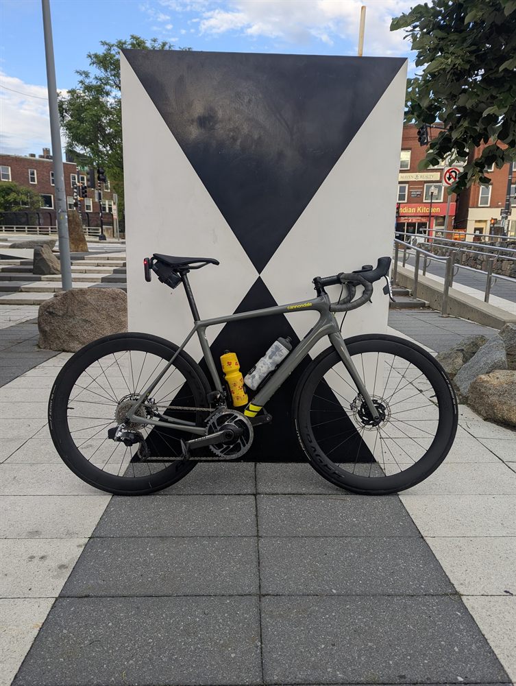 Grey carbon road bike with deep-section wheels, photographed head-on and centred against a large black-and-white geometric sculpture on a paved plaza.