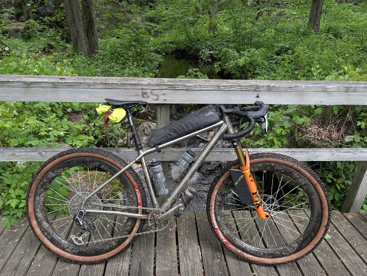 Titanium drop-bar mountain bike with an orange suspension fork and tan-wall knobbly tyres, leaning against the rail of a wooden footbridge over a creek in dense green woods.