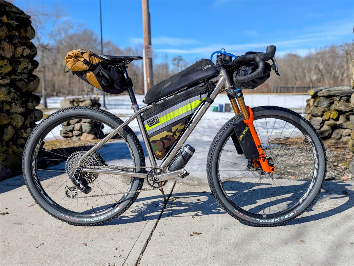 The same titanium drop-bar mountain bike loaded with a bar roll, framebag and seat pack, side on beside a dry stone wall with snow on the ground.