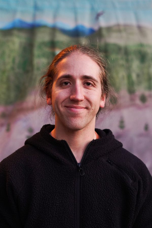 Head-and-shoulders portrait of Jonathan Karpatkin in a black fleece hoodie, warm side light on his face, standing in front of a painted mural of mountains and pine forest.