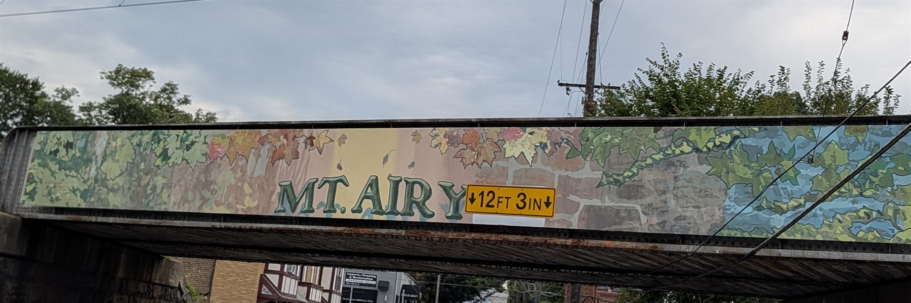 The Mt. Airy railroad bridge, its side painted as a mural of ivy, stone and falling autumn leaves with MT. AIRY lettered across it in green, above a yellow 12 ft 3 in clearance sign and the street running underneath.